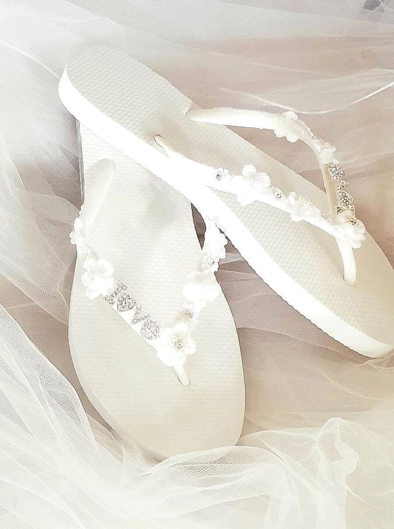 May include: A pair of white flip-flops featuring straps adorned with small white fabric flowers, clear rhinestones, and a silver-toned metal charm that spells out the word love in a cursive script, set against a soft white fabric background.
