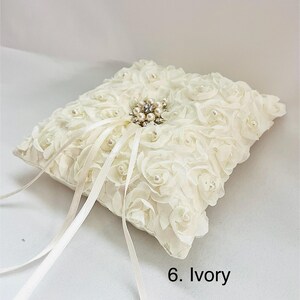 May include: An ivory square ring pillow covered in fabric rosettes and small faux pearls. A central cluster of rhinestones and pearls sits in the middle, with two long satin ribbons extending from the center. The text 6. Ivory is visible.