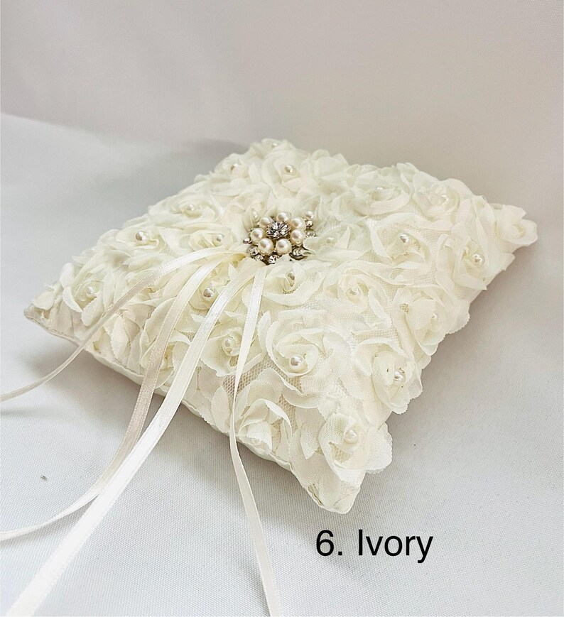 May include: An ivory square ring pillow covered in fabric rosettes and small faux pearls. A central cluster of rhinestones and pearls sits in the middle, with two long satin ribbons extending from the center. The text 6. Ivory is visible.