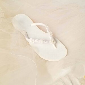 May include: A white flip-flop featuring a textured footbed and a thong strap adorned with small white fabric flowers, tiny white beads, and a single clear rhinestone accent centered on the strap. The sandal is presented in a clean, bright white.