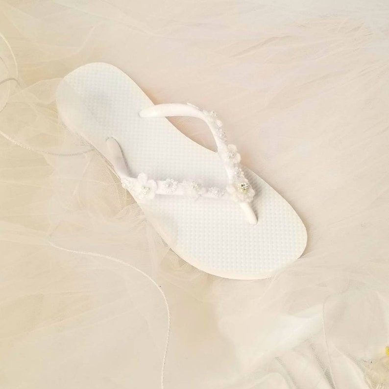 May include: A white flip-flop featuring a textured footbed and a thong strap adorned with small white fabric flowers, tiny white beads, and a single clear rhinestone accent centered on the strap. The sandal is presented in a clean, bright white.