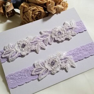 May include: Two lavender garters with white lace and floral embellishments. Each garter has a small purple rhinestone in the center of the flower.