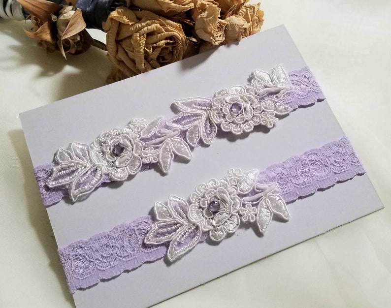May include: Two lavender garters with white lace and floral embellishments. Each garter has a small purple rhinestone in the center of the flower.