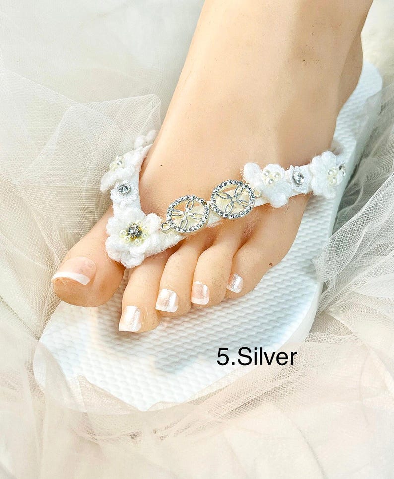 May include: A white flip-flop featuring a strap decorated with white fabric flowers, small pearls, and two circular silver-toned charms with clear crystals. The text 5.Silver is printed on the white textured footbed of the sandal.