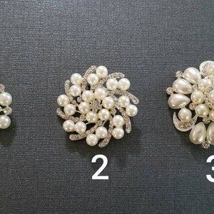 May include: Three silver-toned brooches featuring white faux pearls and clear rhinestones. Brooch 1 is circular with a central pearl. Brooch 2 has a swirling design with pearls and rhinestones. Brooch 3 is a larger floral-shaped piece with teardrop pearls.