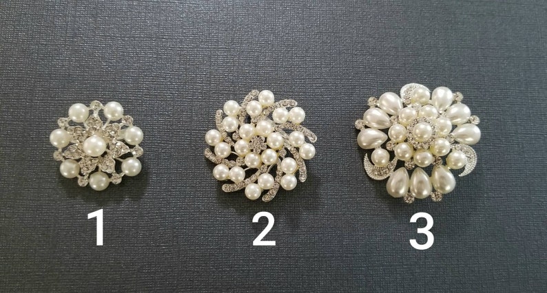 May include: Three silver-toned brooches featuring white faux pearls and clear rhinestones. Brooch 1 is circular with a central pearl. Brooch 2 has a swirling design with pearls and rhinestones. Brooch 3 is a larger floral-shaped piece with teardrop pearls.