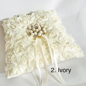 May include: An ivory square ring pillow covered in fabric rosettes with small pearls at their centers. A central brooch features a cluster of pearls and clear crystals. Two long satin ribbons extend from the center. The text 2. Ivory is visible.