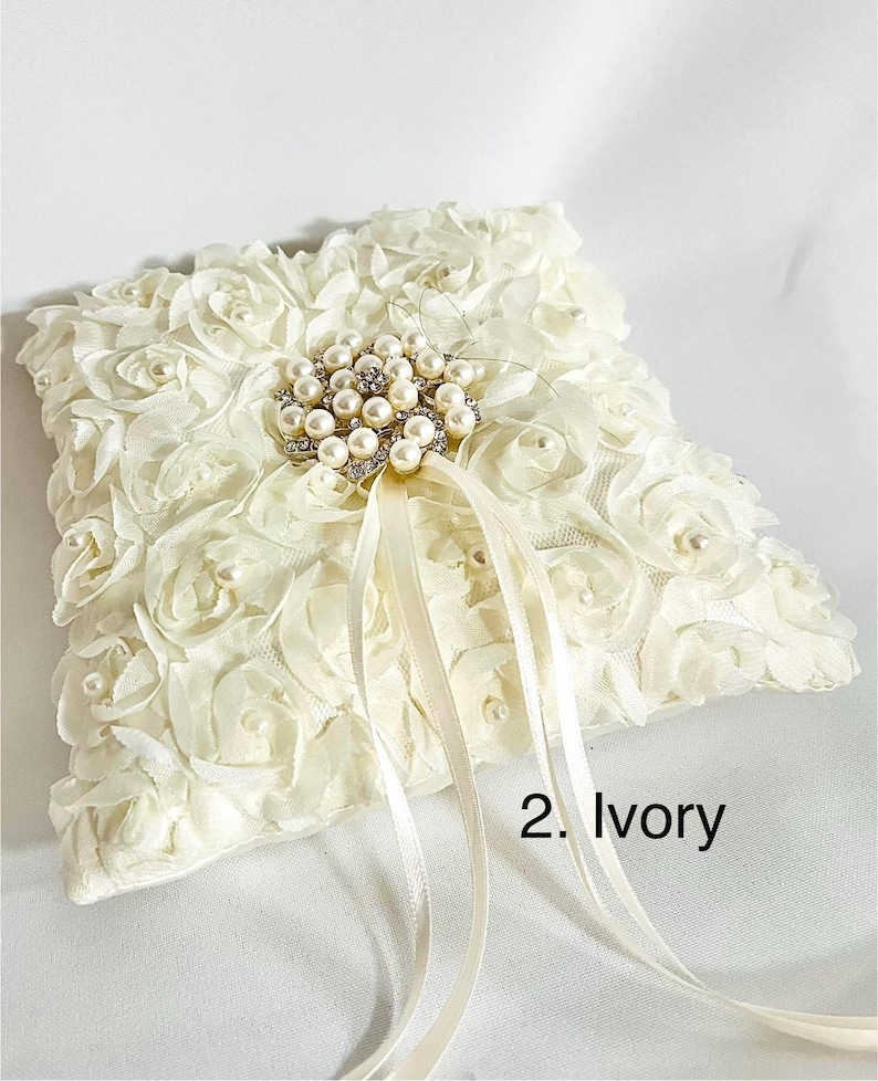 May include: An ivory square ring pillow covered in fabric rosettes with small pearls at their centers. A central brooch features a cluster of pearls and clear crystals. Two long satin ribbons extend from the center. The text 2. Ivory is visible.