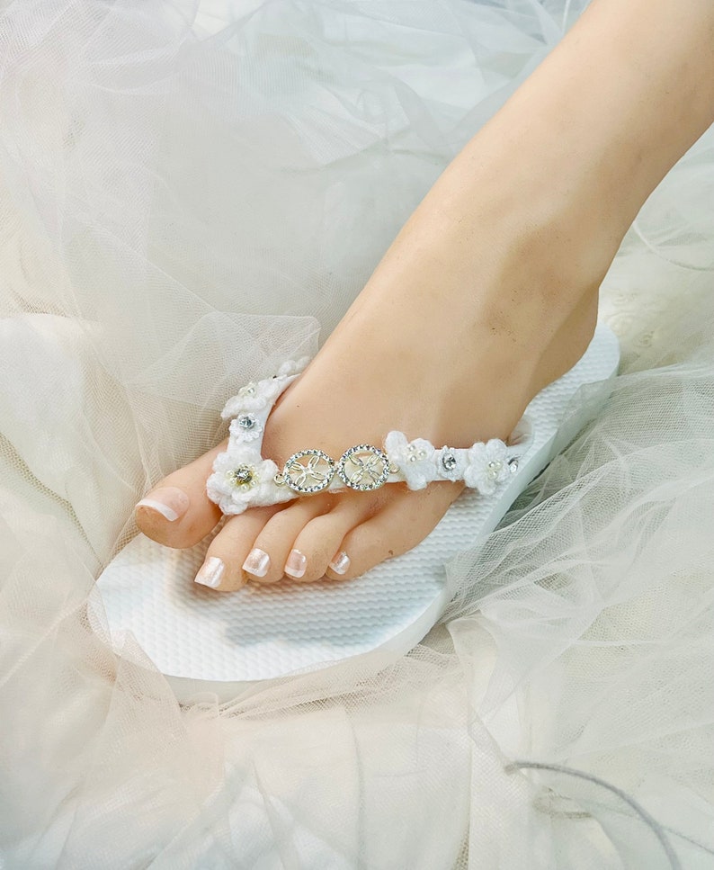 May include: A white flip-flop sandal featuring a textured sole and a strap adorned with white crocheted flowers, small faux pearls, and two circular silver-toned metal charms embedded with clear rhinestones arranged in a horizontal row.