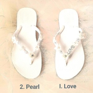 May include: A pair of white flip-flops. The left sandal, labeled 2. Pearl, features white floral and pearl embellishments on the straps. The right sandal, labeled 1. Love, features floral accents and silver-toned letters spelling out the word LOVE.