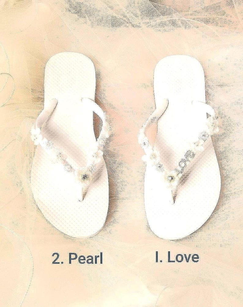 May include: A pair of white flip-flops. The left sandal, labeled 2. Pearl, features white floral and pearl embellishments on the straps. The right sandal, labeled 1. Love, features floral accents and silver-toned letters spelling out the word LOVE.