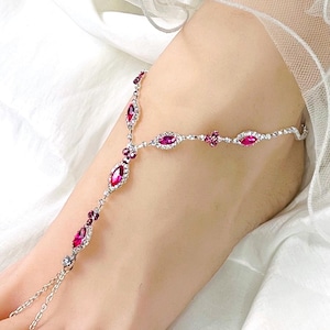 May include: A silver foot chain with ruby-colored gemstones and clear crystals. The anklet features a delicate design with a central pendant and a chain that connects to the toe. The gemstones are oval and round, set in a silver metal.