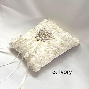 May include: An ivory ring pillow covered in fabric roses with small pearls at their centers. A central brooch features clusters of pearls and clear crystals. Two long, thin satin ribbons extend from the center. The text 3. Ivory is visible.