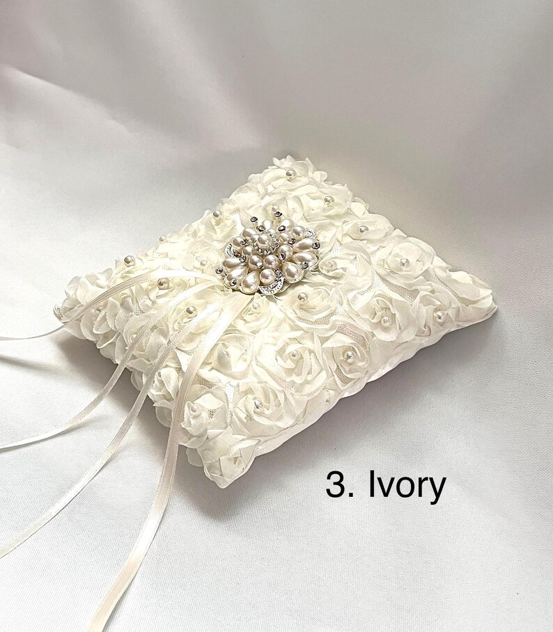 May include: An ivory ring pillow covered in fabric roses with small pearls at their centers. A central brooch features clusters of pearls and clear crystals. Two long, thin satin ribbons extend from the center. The text 3. Ivory is visible.