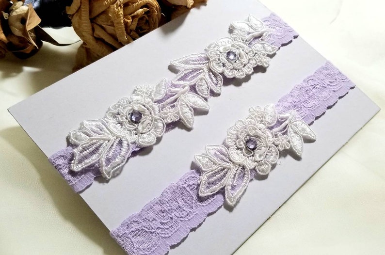May include: Two white lace garters with purple trim and a rhinestone embellishment on each. The garters are displayed on a white background.