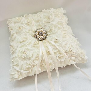 May include: A square ivory ring pillow covered in fabric rosettes and small white faux pearls. A central silver-toned brooch features a cluster of clear crystals and faux pearls, with two long, thin ivory satin ribbons extending downward.