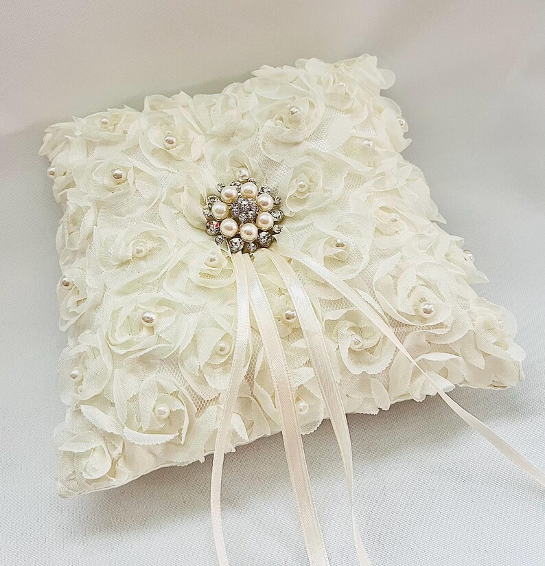 May include: A square ivory ring pillow covered in fabric rosettes and small white faux pearls. A central silver-toned brooch features a cluster of clear crystals and faux pearls, with two long, thin ivory satin ribbons extending downward.