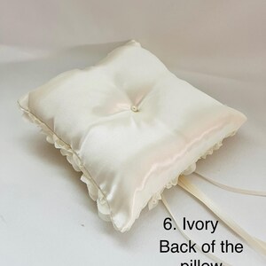 May include: A square ivory satin ring pillow featuring a central faux pearl accent, ruffled fabric trim along the edges, and long satin ribbons attached to the corner. The text 6. Ivory Back of the pillow is printed in black on the lower right side.