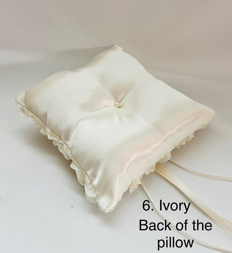 May include: A square ivory satin ring pillow featuring a central faux pearl accent, ruffled fabric trim along the edges, and long satin ribbons attached to the corner. The text 6. Ivory Back of the pillow is printed in black on the lower right side.