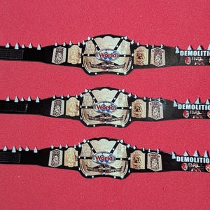 May include: Three black wrestling championship belts featuring silver-toned centre plates with the text World Tag Team Wrestling Champions. The straps include white spikes along the top edge and the word Demolition printed in white on the right side.