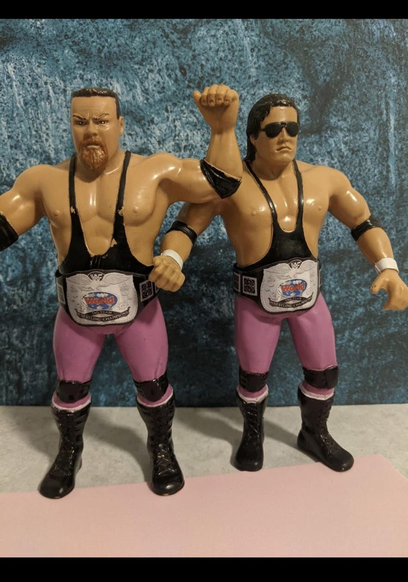 Pu&ograve; includere: Due action figure di lottatori. Indossano canottiere nere, pantaloni rosa e stivali neri. Ogni figura ha una cintura da campione. Una figura ha la barba e l'altra indossa occhiali da sole. Le figure sono in posa con un braccio alzato.