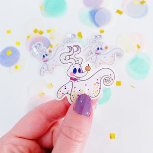 Könnte beinhalten: Ein weißer Geistercharakter mit rosa Nase und blauen Augen, der einen kleinen orangefarbenen Kürbis hält. Der Geist ist von einem weißen Hintergrund mit goldenem und blauem Glitzer umgeben.