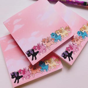 Könnte beinhalten: Rosa quadratische Notizblöcke mit einem Himmelshintergrund und einer Reihe von bunten geflügelten Ponys am unteren Rand. Die Ponys haben verschiedene Farben, darunter Schwarz, Rosa, Blau und Gelb.