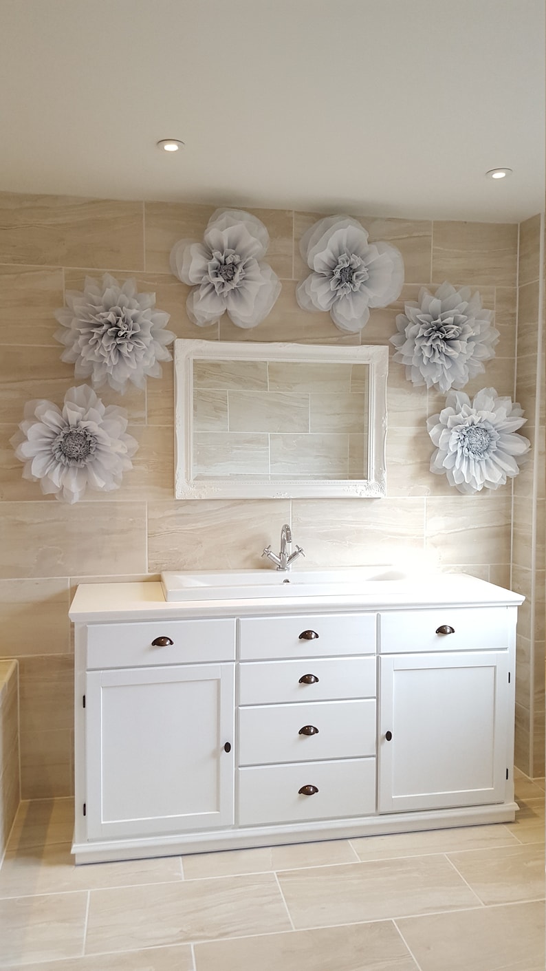 Puede incluir: Mueble de ba&ntilde;o blanco con un lavabo de porcelana blanca y un grifo cromado. El mueble tiene seis cajones y dos puertas. La pared detr&aacute;s del mueble est&aacute; alicatada en un color beige claro y decorada con seis grandes flores de papel grises.