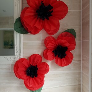 May include: Three large red paper poppies with black centers and green leaves are displayed on a tiled wall.
