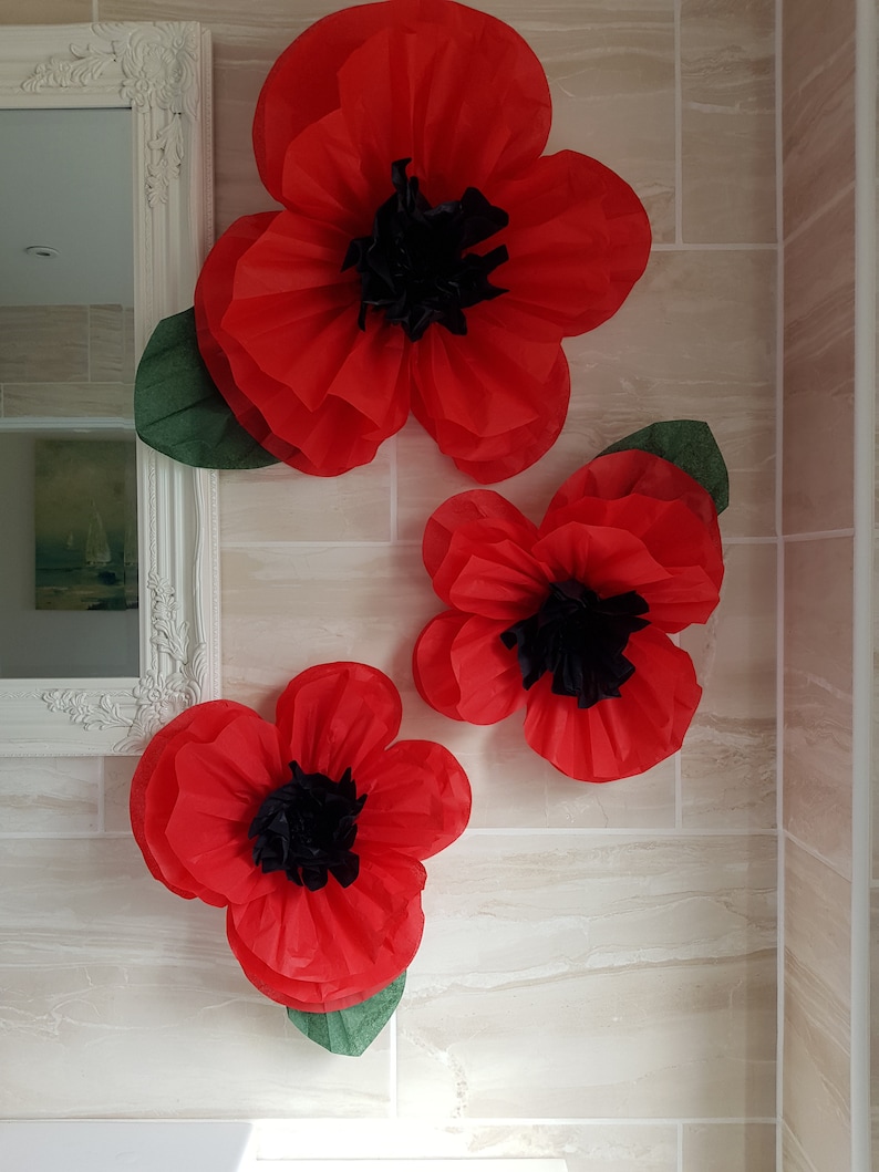 May include: Three large red paper poppies with black centers and green leaves are displayed on a tiled wall.