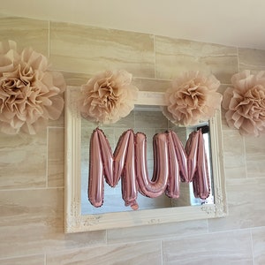 Puede incluir: Un espejo con marco blanco con un globo de aluminio rosa que deletrea "MAM&Aacute;" en el reflejo. El espejo est&aacute; decorado con cuatro grandes pompones de papel beige.
