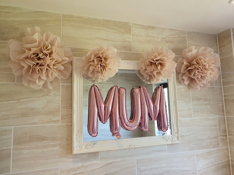 Puede incluir: Un espejo con marco blanco con un globo de aluminio rosa que deletrea "MAM&Aacute;" en el reflejo. El espejo est&aacute; decorado con cuatro grandes pompones de papel beige.