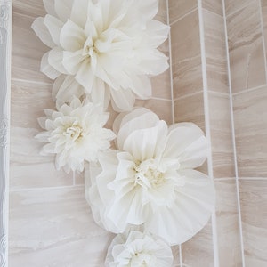 Puede incluir: Cuatro grandes flores de papel blanco est&aacute;n dispuestas en una pared de azulejos beige. Las flores est&aacute;n hechas de papel de seda y tienen una textura delicada y arrugada.