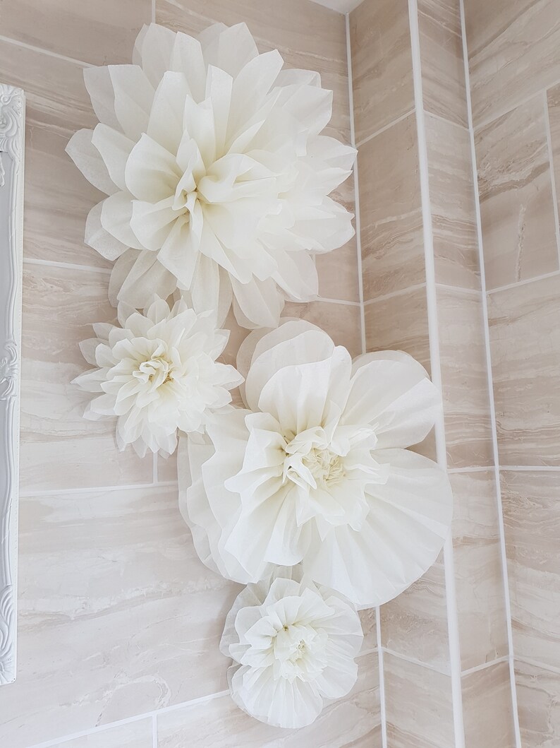 Puede incluir: Cuatro grandes flores de papel blanco est&aacute;n dispuestas en una pared de azulejos beige. Las flores est&aacute;n hechas de papel de seda y tienen una textura delicada y arrugada.