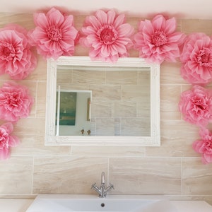 May include: A bathroom decorated with pink paper flowers. The flowers are arranged around a white framed mirror and on the wall. A white porcelain sink with a chrome faucet is in the foreground.