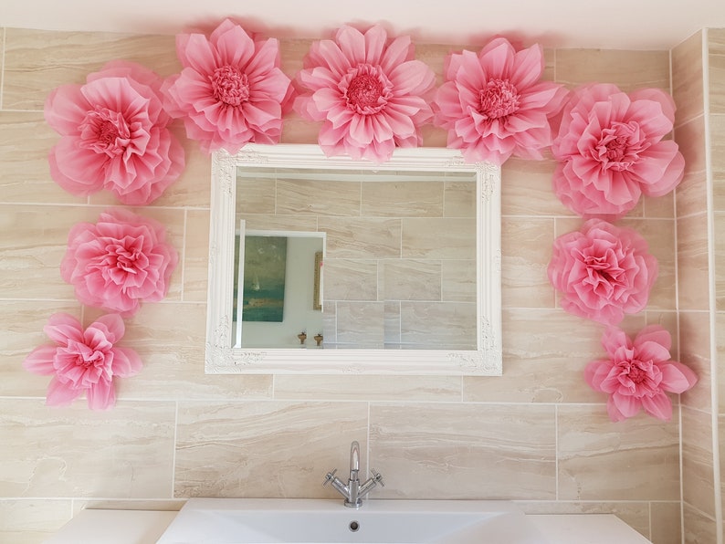 May include: A bathroom decorated with pink paper flowers. The flowers are arranged around a white framed mirror and on the wall. A white porcelain sink with a chrome faucet is in the foreground.