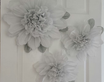 May include: Three large gray paper flowers are attached to a white door. The flowers are made of tissue paper and have a layered, ruffled appearance.