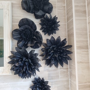 Puede incluir: Un conjunto de seis flores de papel negro, cada una con una forma y un tama&ntilde;o diferentes, dispuestas en una pared de azulejos beige. Las flores est&aacute;n hechas de papel de seda y tienen un aspecto delicado y en capas.