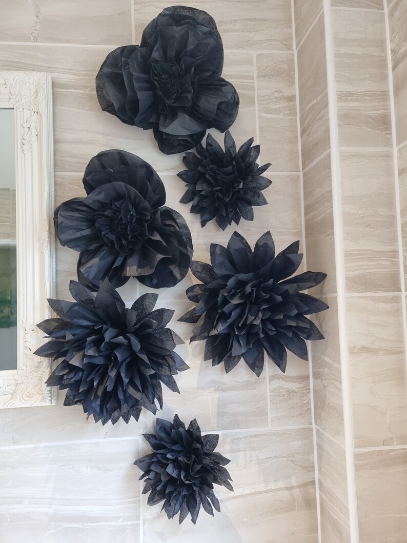 Puede incluir: Un conjunto de seis flores de papel negro, cada una con una forma y un tama&ntilde;o diferentes, dispuestas en una pared de azulejos beige. Las flores est&aacute;n hechas de papel de seda y tienen un aspecto delicado y en capas.