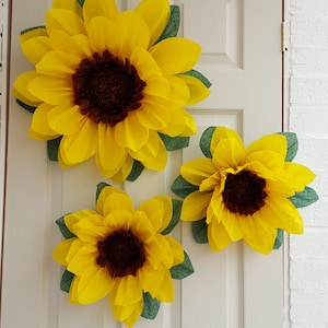 May include: Three large paper sunflowers with brown centers and green leaves are attached to a white door.
