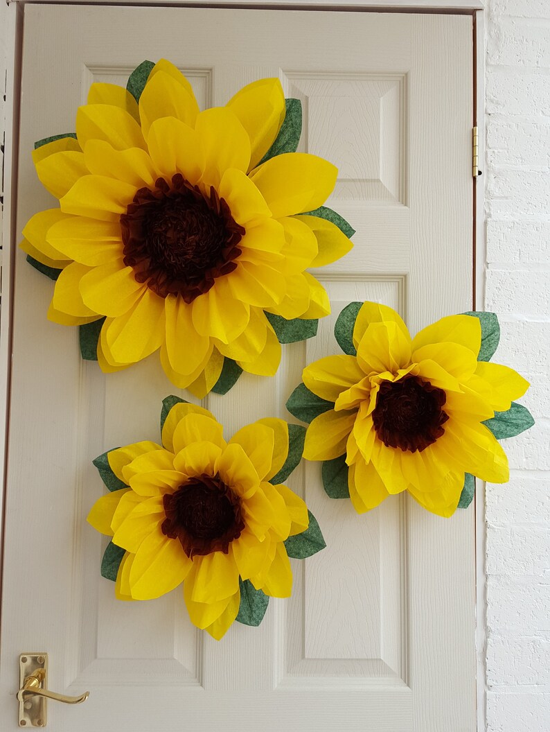 May include: Three large paper sunflowers with brown centers and green leaves are attached to a white door.