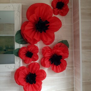 Puede incluir: Cinco amapolas de papel rojo con centros negros est&aacute;n dispuestas en una pared de azulejos blancos. Las amapolas est&aacute;n superpuestas para crear un efecto 3D.