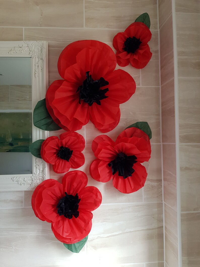Puede incluir: Cinco amapolas de papel rojo con centros negros est&aacute;n dispuestas en una pared de azulejos blancos. Las amapolas est&aacute;n superpuestas para crear un efecto 3D.