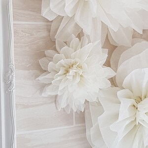 Puede incluir: Flores de papel de seda blancas en varios tama&ntilde;os y formas. Las flores est&aacute;n dispuestas en una pared blanca con un patr&oacute;n de azulejos de color marr&oacute;n claro.