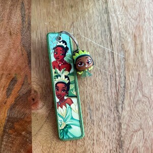 May include: A green and white keychain featuring images of Princess Tiana from the movie "The Princess and the Frog." The keychain has a small, attached figurine of Princess Tiana. The background is a wooden surface.