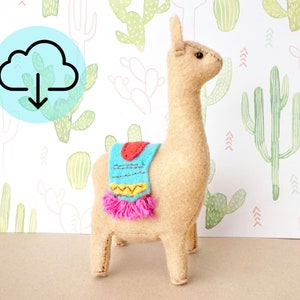 Könnte beinhalten: Ein beigefarbener Filz-Lama mit einem blau, gelb und pink gemusterten Sattel. Der Lama steht auf einem weißen Hintergrund mit einem grünen Kaktus-Muster.