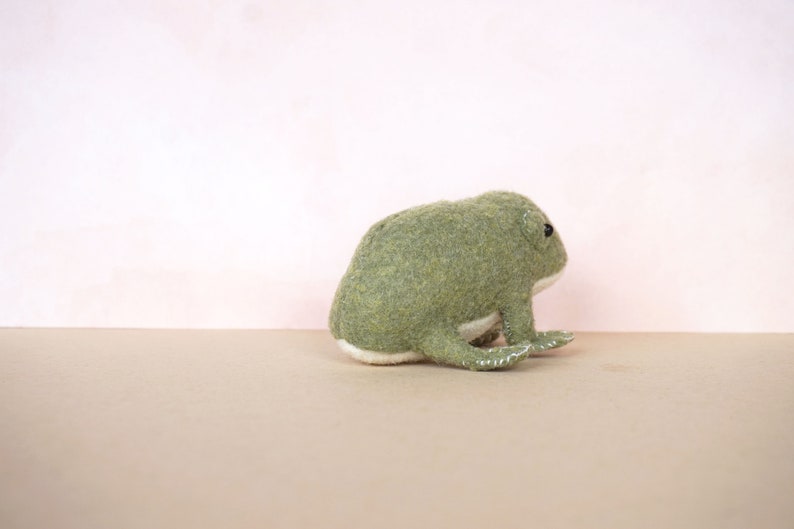 May include: A green felt frog plush toy sitting on a light brown surface. The frog has a white belly and black eyes.