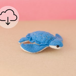 May include: A small blue felt turtle with white accents and black eyes. The turtle is lying on a light brown surface.