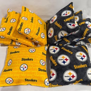 May include: A collection of bean bags in yellow and black fabric, featuring the Pittsburgh Steelers logo and team name. The bean bags are square and triangular, suitable for a game of cornhole or similar activities.