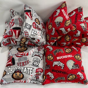 May include: Six bean bags with a grey and red fabric design. The bean bags feature the Ohio State Buckeyes logo and mascot, Brutus Buckeye. The bean bags are arranged in a pile.